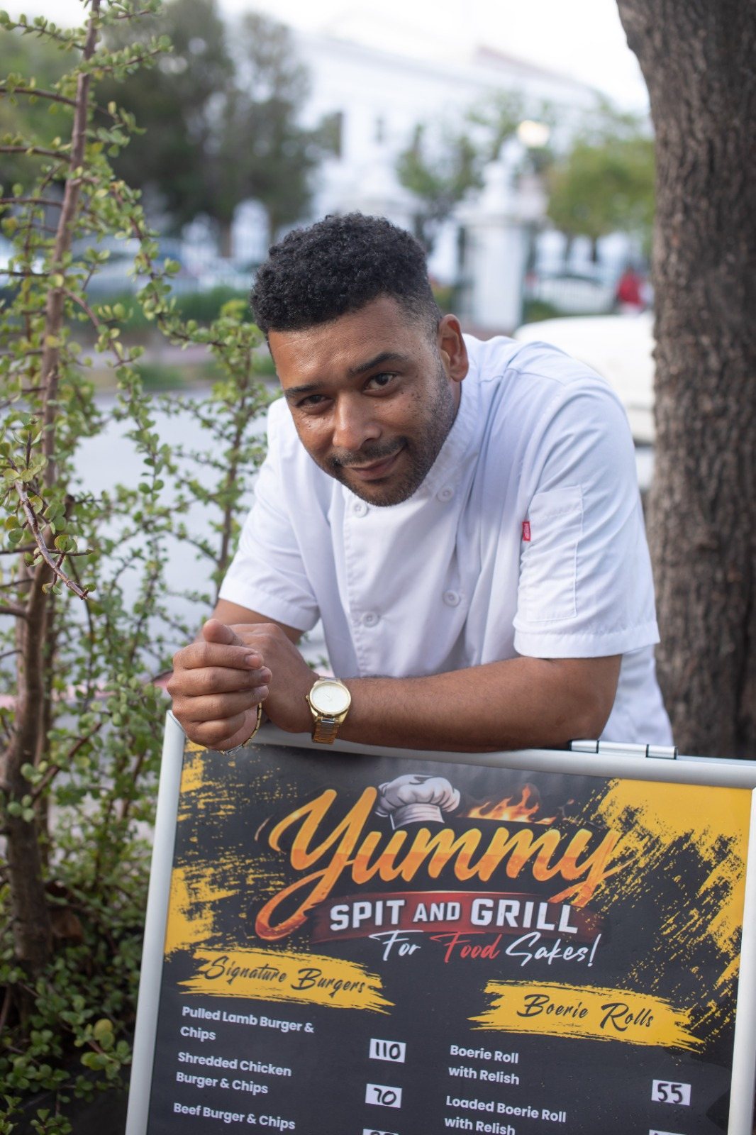 A Yummy Spit & Grill chef leaning on the menu board outside the shop