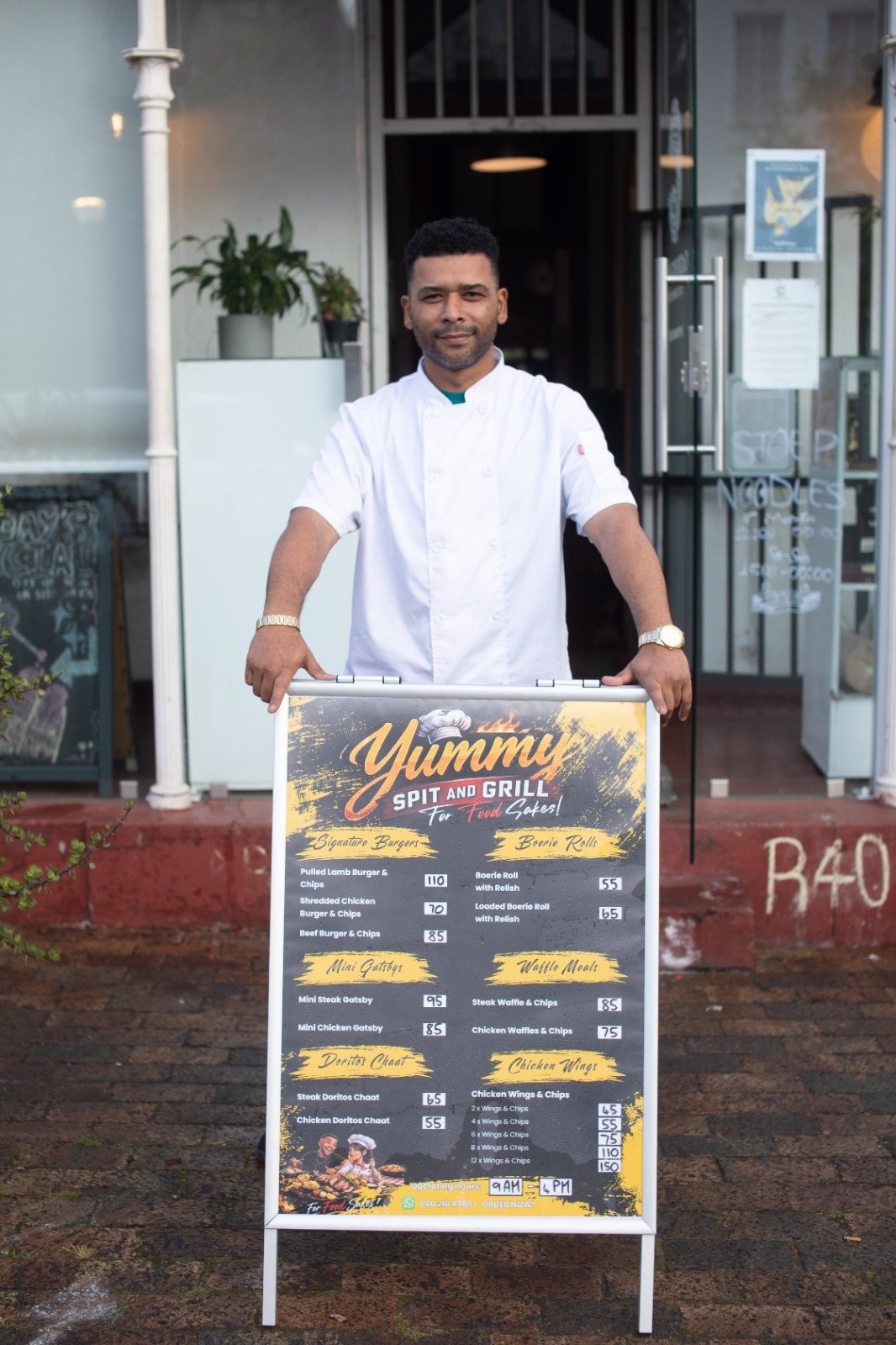 A Yummy Spit & Grill chef standing behind the menu board outside the shop