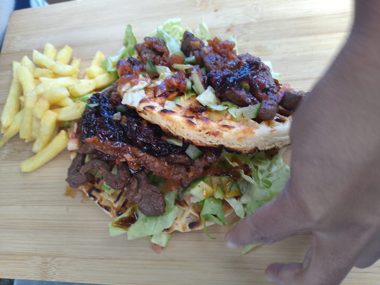 A hand steadying a board of grilled steak on a crispy waffle with chips